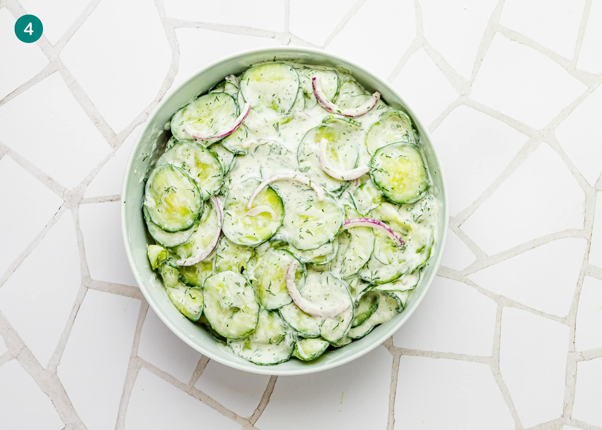 A bowl of Creamy Cucumber Salad (Egg-Free) with sliced red onions and herbs sits on a white tiled surface.