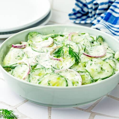 A bowl of Creamy Cucumber Salad (Egg-Free) with sliced cucumbers, red onions, and fresh dill sits on a white tiled surface next to plates and a blue striped towel.