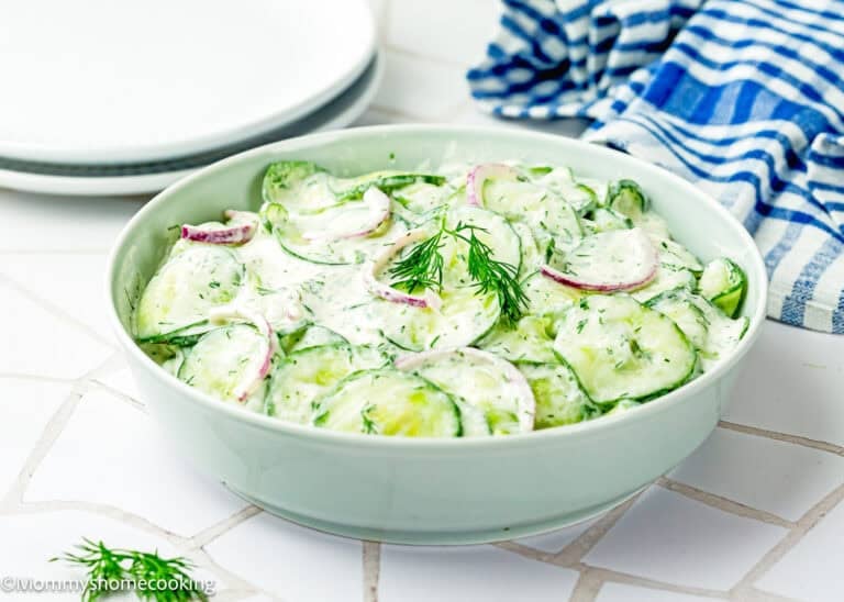 A bowl of Creamy Cucumber Salad (Egg-Free) with sliced cucumbers, red onions, and fresh dill sits on a white tiled surface next to plates and a blue striped towel.