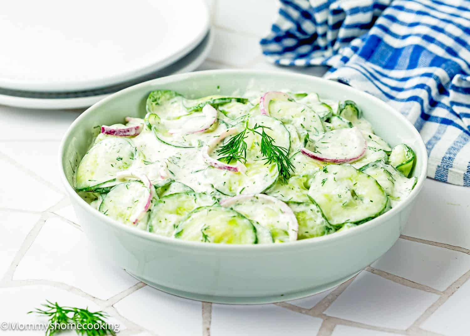 A bowl of Creamy Cucumber Salad (Egg-Free) with sliced cucumbers, red onions, and fresh dill sits on a white tiled surface next to plates and a blue striped towel.