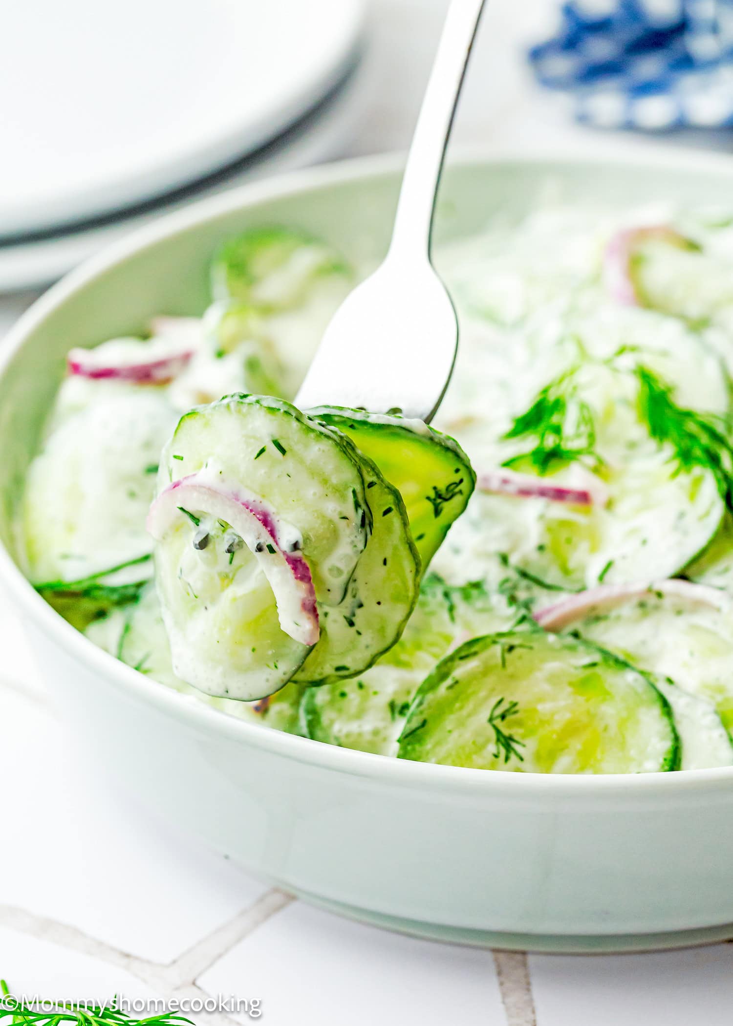 A fork holds sliced cucumber and red onion coated in a creamy dressing, above a bowl of Creamy Cucumber Salad (Egg-Free) garnished with fresh dill.