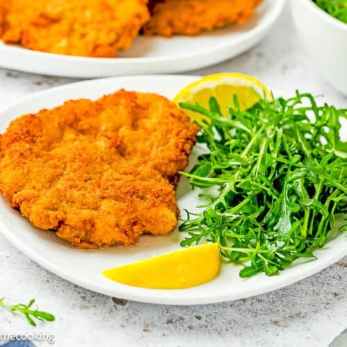A plate with Crispy Eggless Pork Schnitzel (egg and dairy free), a serving of fresh arugula, and two lemon wedges.