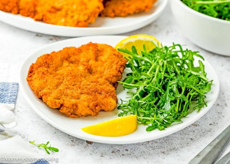 A plate with Crispy Eggless Pork Schnitzel (egg and dairy free), a serving of fresh arugula, and two lemon wedges.
