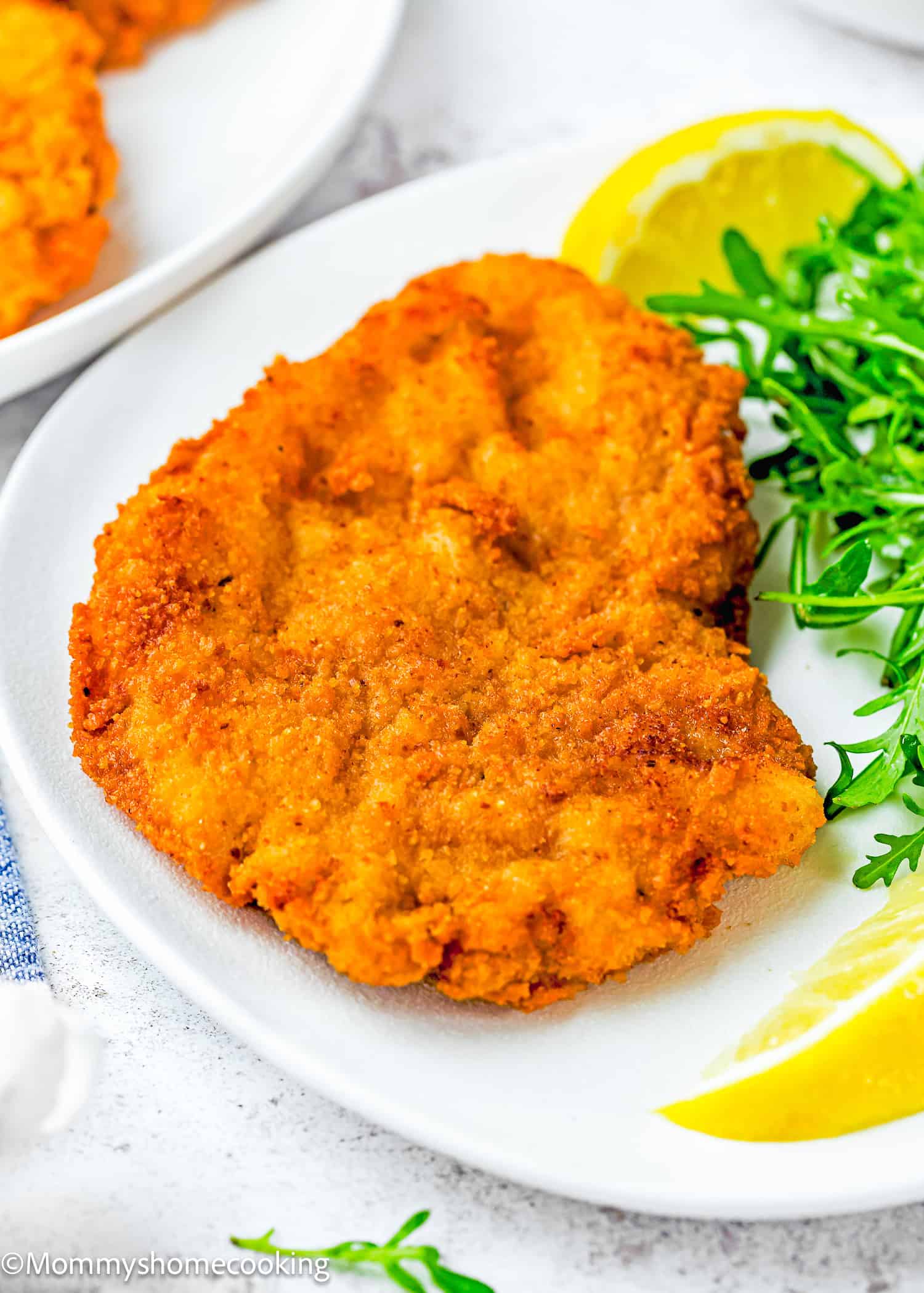A crispy, eggless pork schnitzel (egg and dairy free) is served on a white plate with fresh greens and lemon wedges.