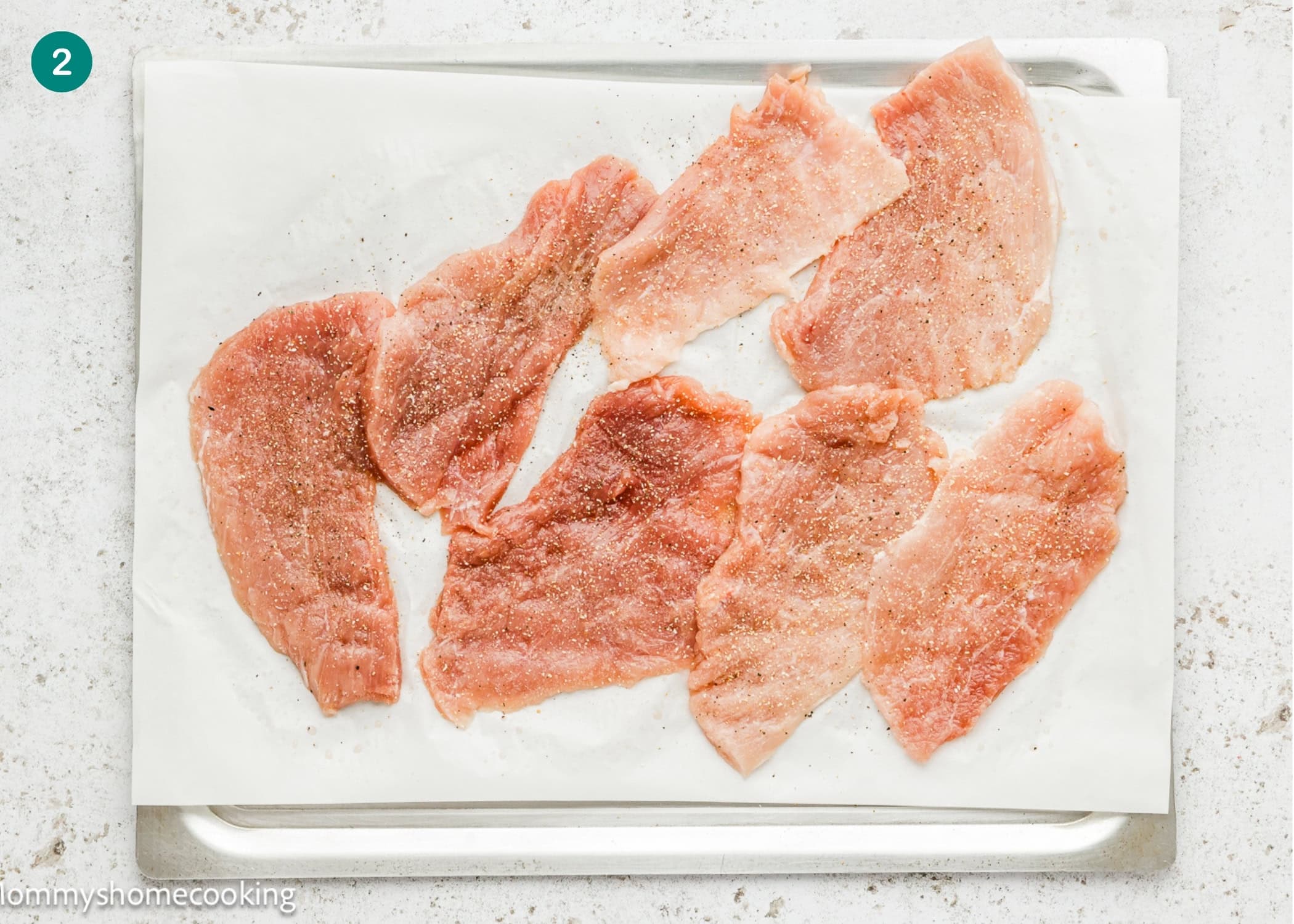 Raw, thinly sliced pieces of meat are spread out on a baking sheet lined with parchment paper, lightly seasoned with salt and pepper for a crispy eggless pork schnitzel (egg and dairy free).