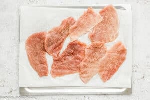 Raw, thinly sliced pieces of meat are spread out on a baking sheet lined with parchment paper, lightly seasoned with salt and pepper for a crispy eggless pork schnitzel (egg and dairy free).