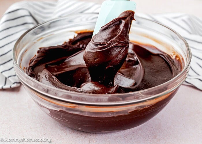 A glass bowl filled with smooth Easy Chocolate Fudge Frosting being stirred with a blue spatula, with a striped cloth in the background.