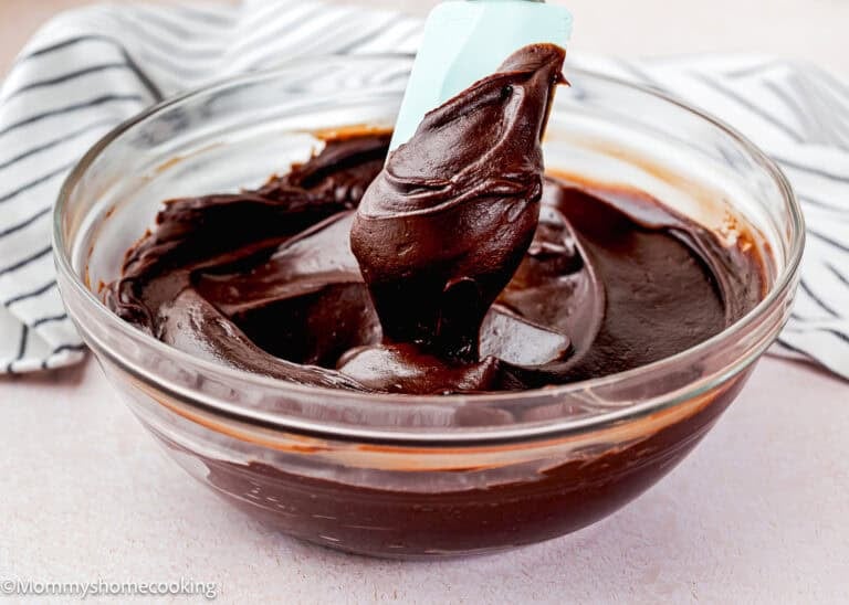 A glass bowl filled with smooth Easy Chocolate Fudge Frosting being stirred with a blue spatula, with a striped cloth in the background.