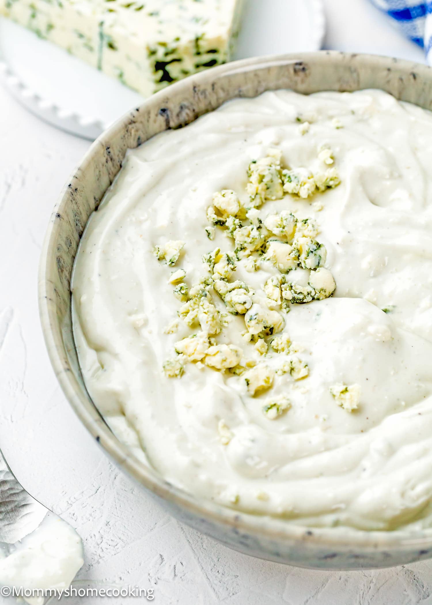 A bowl of creamy Easy Eggless Blue Cheese Dressing topped with crumbled blue cheese, with a wedge of blue cheese in the background.