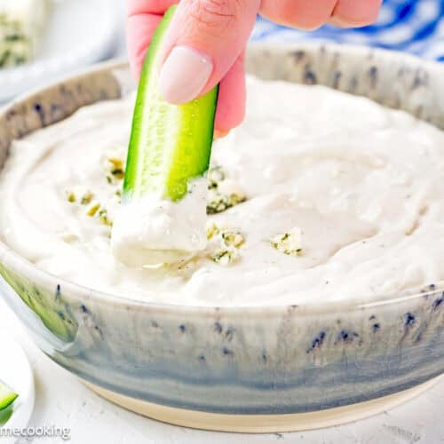 A hand dips a cucumber stick into a bowl of Easy Eggless Blue Cheese Dressing, with more cucumber sticks on the side.