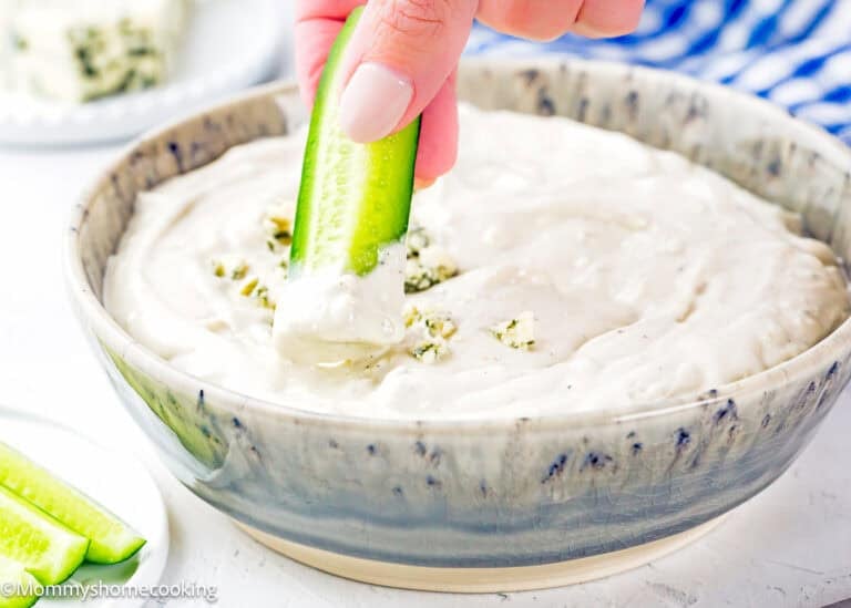 A hand dips a cucumber stick into a bowl of Easy Eggless Blue Cheese Dressing, with more cucumber sticks on the side.