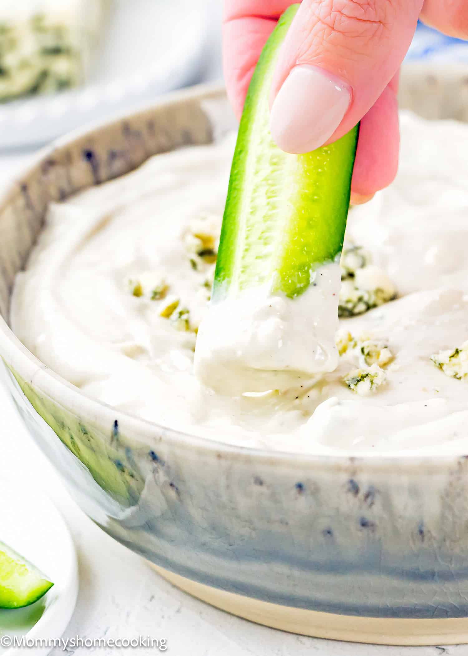A hand dips a cucumber slice into a bowl of creamy Easy Eggless Blue Cheese Dressing topped with fresh herbs.