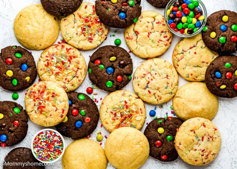 Easy Eggless Cake Mix Cookies with colorful sprinkles and candy-coated chocolates are arranged on a white surface, alongside two small bowls of extra sprinkles and candies.