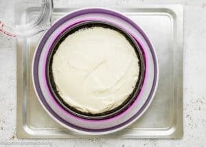 An Eggless Baileys Irish Cream Cheesecake (Baked) with an Oreo crust in a round pan sits inside a purple silicone ring on a metal tray, with a glass measuring cup nearby.