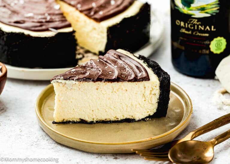 A slice of Eggless Baileys Irish Cream Cheesecake (Baked) with a chocolate topping and dark crust sits on a plate, accompanied by a fork, spoon, and a bottle of Irish cream in the background.