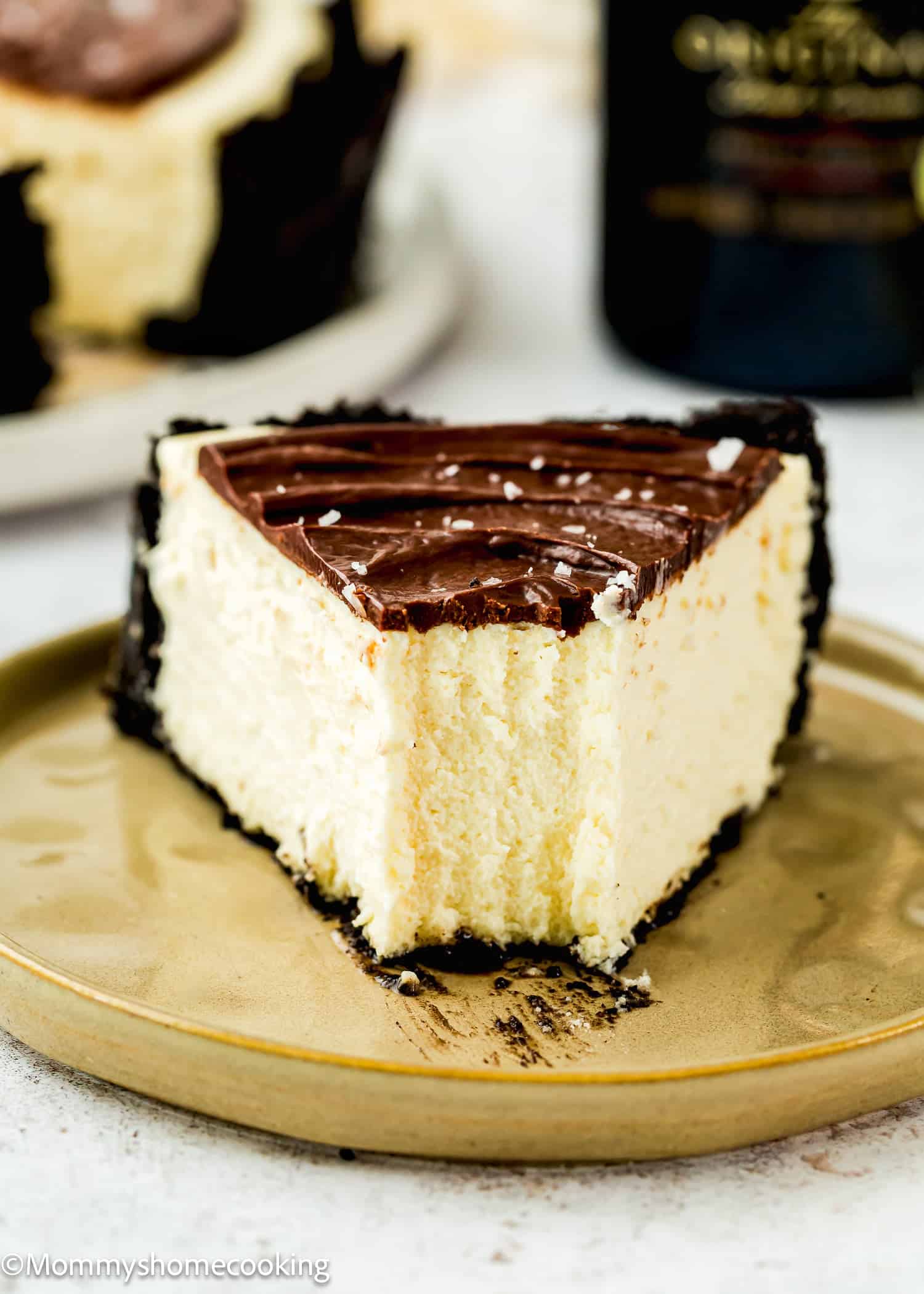 A slice of Eggless Baileys Irish Cream Cheesecake (Baked) with chocolate topping and an Oreo crust sits on a beige plate, with a bite taken out of it.