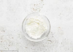 A clear glass bowl filled with white ricotta cheese, perfect for making Eggless Baileys Irish Cream Cheesecake (Baked), sits on a textured white surface.