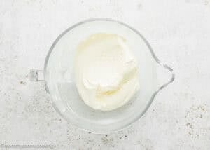 A clear glass bowl filled with thick, creamy white Eggless Baileys Irish Cream Cheesecake (Baked) mixture sits on a light, speckled surface.