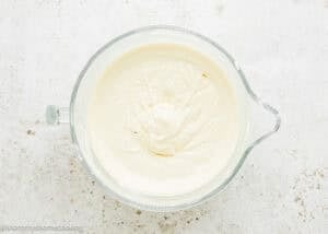 A glass mixing bowl filled with smooth, creamy white Eggless Baileys Irish Cream Cheesecake (Baked) batter on a light textured surface.