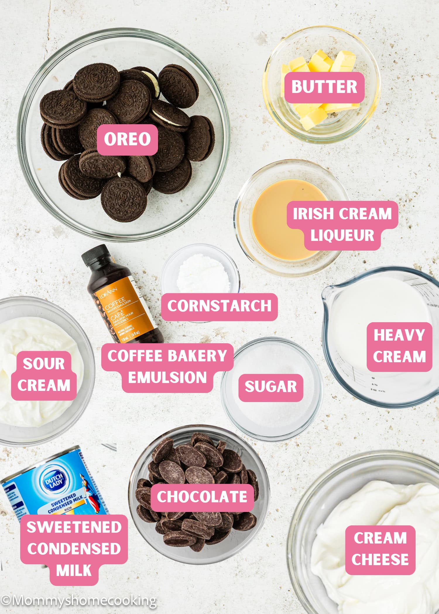 Overhead view of labeled Eggless Baileys Irish Cream Cheesecake (Baked) ingredients in bowls, including Oreo cookies, butter, Irish cream liqueur, cornstarch, coffee bakery emulsion, sugar, heavy cream, chocolate, sour cream, condensed milk, and cream cheese.