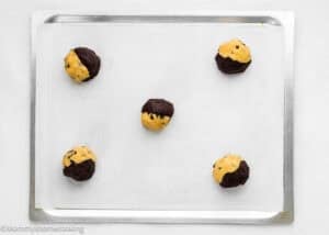 A baking sheet with five Eggless Brookie Cookies (One Dough), each cookie dough ball half chocolate and half plain, spaced apart on parchment paper.