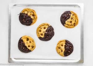 Five half-chocolate, half-vanilla Eggless Brookie Cookies (One Dough) with chocolate chips on a parchment-lined baking sheet.