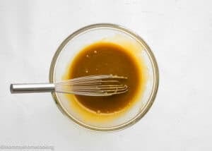 A glass bowl filled with a brown liquid mixture for Eggless Brookie Cookies (One Dough) and a metal whisk on a white surface.