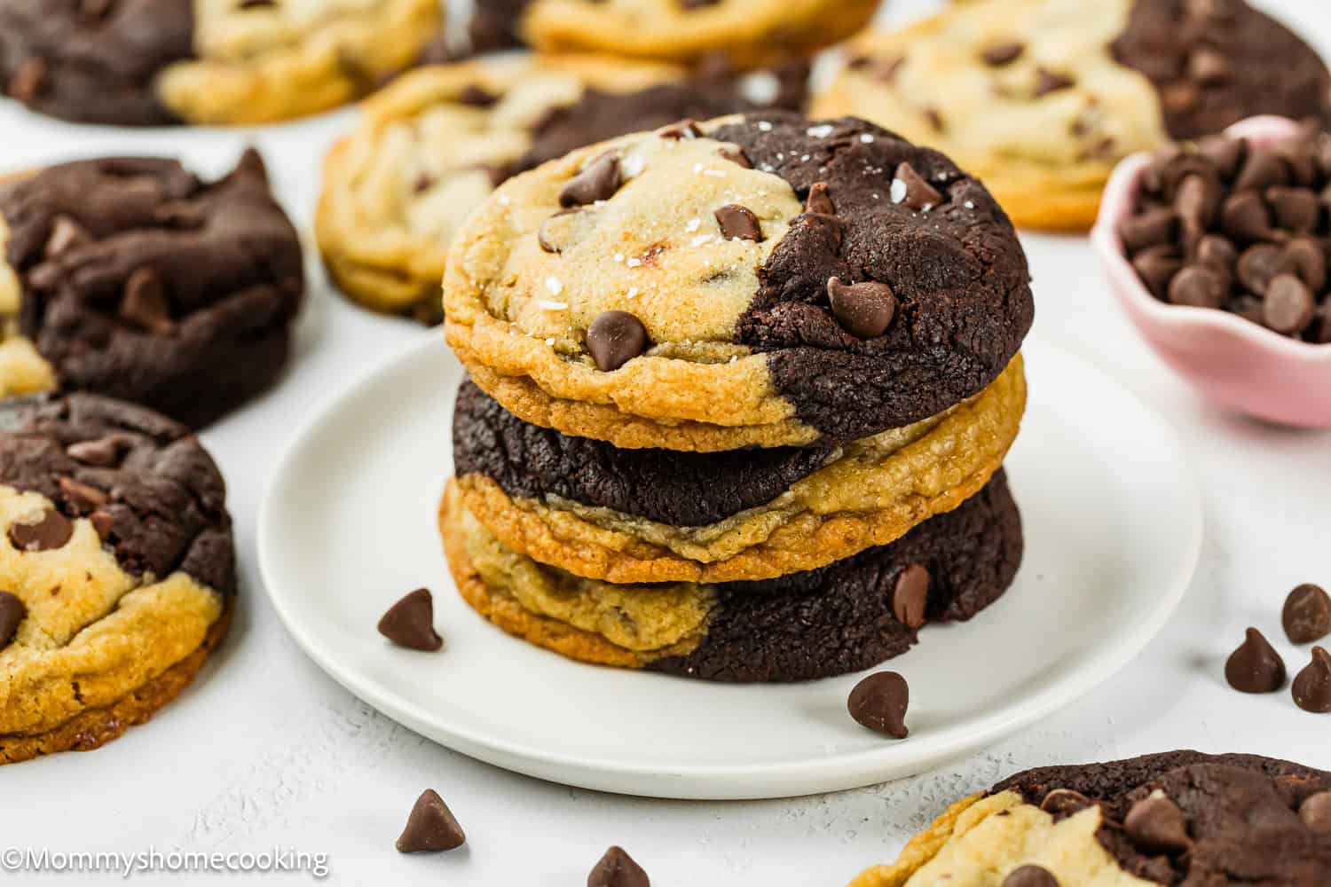 A plate of stacked Eggless Brookie Cookies (One Dough) with half chocolate and half vanilla dough, surrounded by more cookies and scattered chocolate chips.