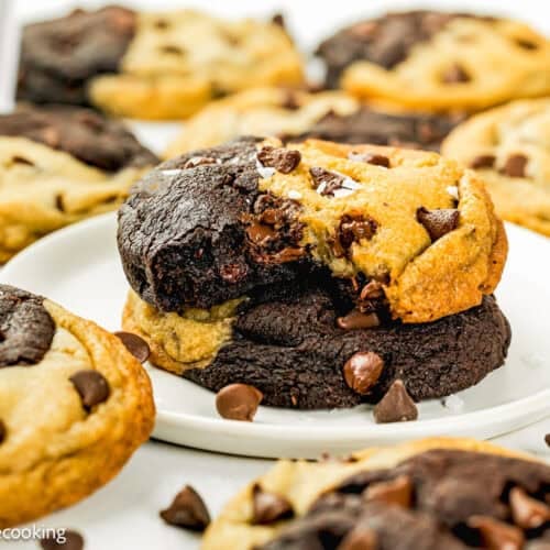 A plate of Eggless Brookie Cookies (One Dough), half-chocolate and half-vanilla with chocolate chips, is surrounded by more cookies and scattered chocolate chips.