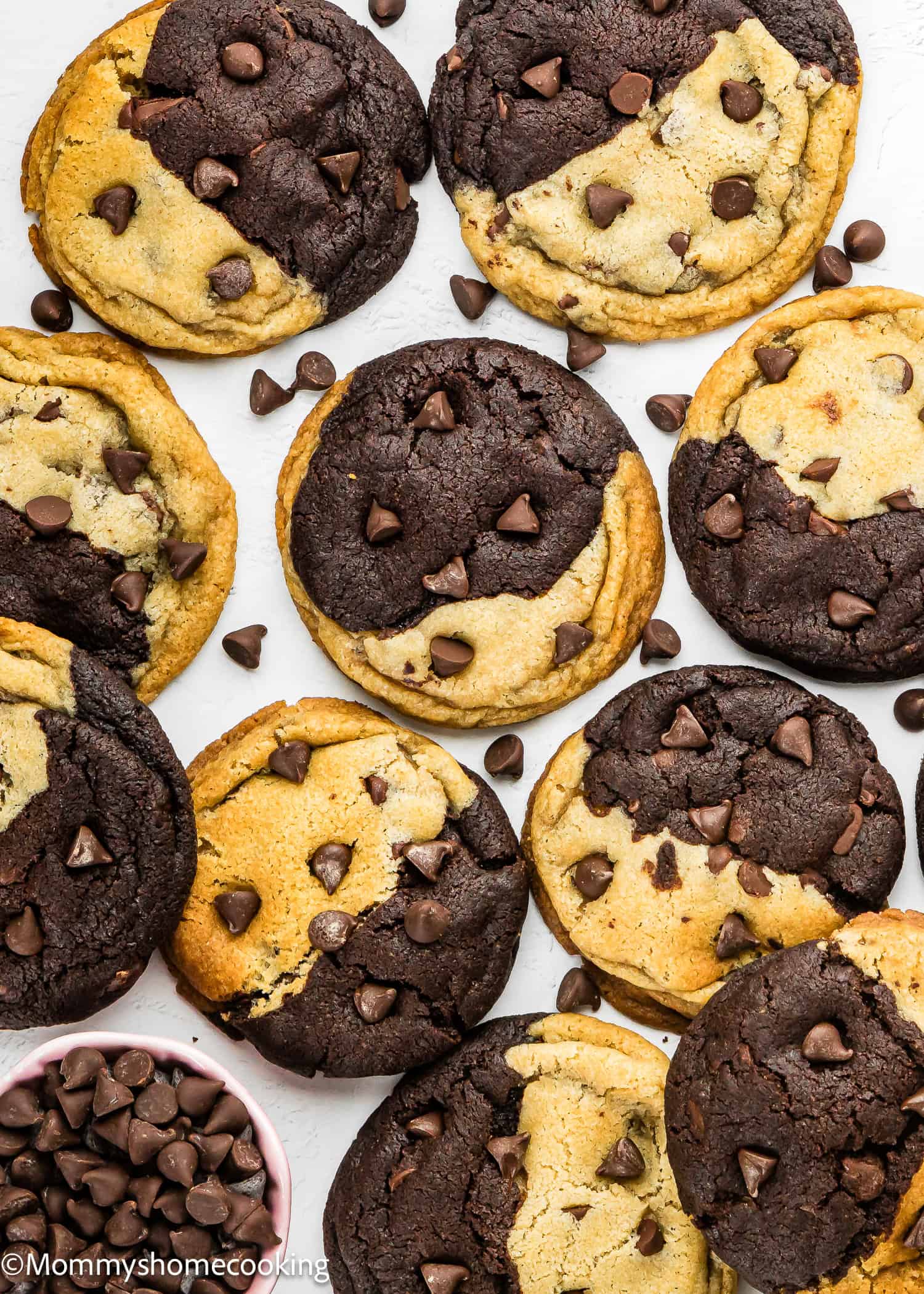 A batch of Eggless Brookie Cookies (One Dough) with chocolate and vanilla swirls and chocolate chips, arranged on a white surface beside a small bowl of chocolate chips.