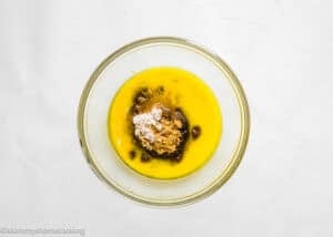 A glass bowl contains melted butter, brown sugar, and flour on a white surface, ready to be mixed for eggless brookie cookies (one dough).