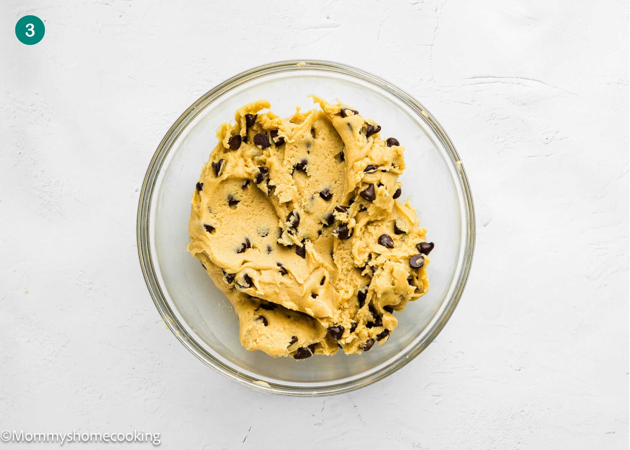A glass bowl filled with eggless Brookie Cookies (one dough) chocolate chip cookie dough sits on a white surface.