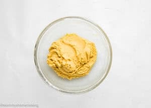 A glass bowl filled with a thick, yellowish dough mixture for Eggless Brookie Cookies (One Dough) sits on a white surface.
