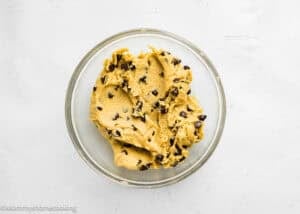A glass bowl filled with eggless brookie cookies (one dough) chocolate chip cookie dough on a white surface.