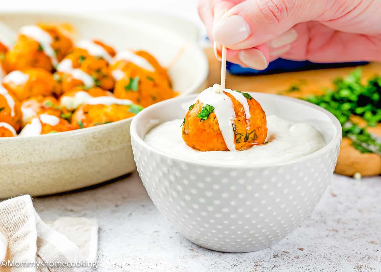 A hand dips a toothpick-skewered Eggless Buffalo Chicken Meatball with sauce into a bowl of creamy dip, with more meatballs and chopped herbs in the background.