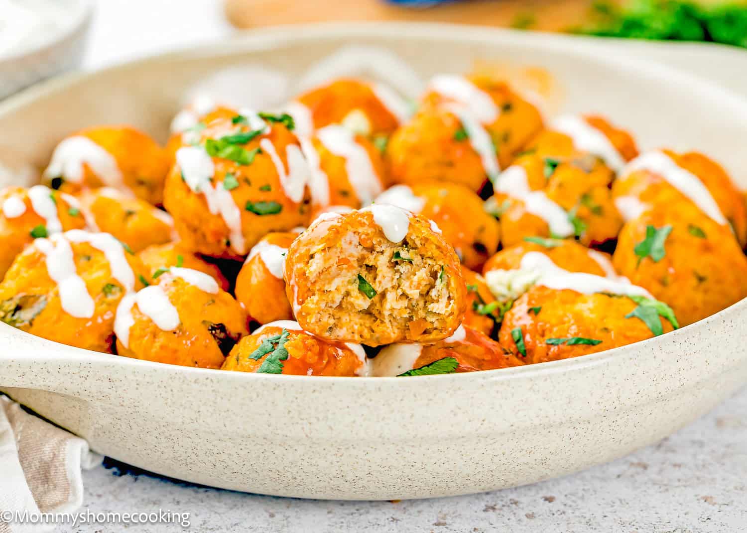 A shallow bowl filled with Eggless Buffalo Chicken Meatballs glazed in orange sauce, garnished with chopped herbs and drizzled with white sauce; one meatball is cut in half to show the inside.