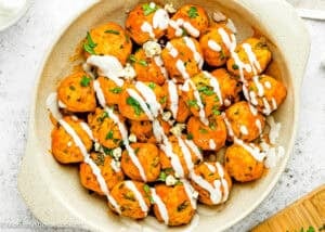 A dish of Eggless Buffalo Chicken Meatballs topped with white sauce, chopped herbs, and crumbled cheese in a beige baking dish.