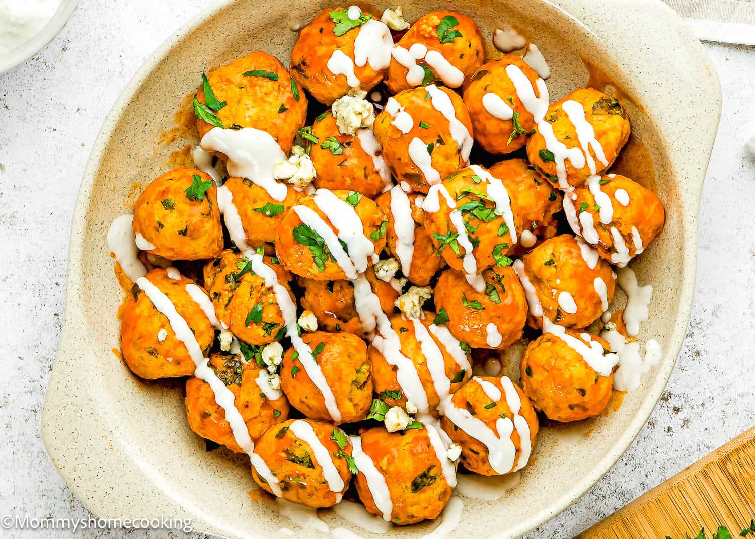 A dish of Eggless Buffalo Chicken Meatballs topped with white sauce, chopped herbs, and crumbled cheese in a beige baking dish.