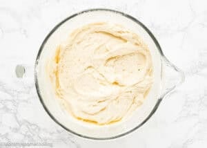 A glass mixing bowl filled with creamy, pale yellow Eggless Marble Bundt Cake batter on a white marble surface.