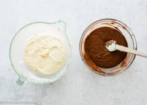 Two glass bowls on a white surface: one with vanilla cake batter, the other with chocolate batter and a spatula, ready for making an Eggless Marble Bundt Cake.