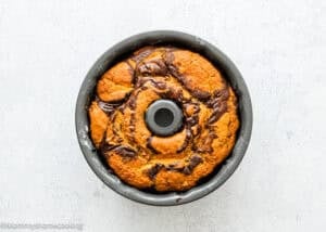 An Eggless Marble Bundt Cake with a chocolate and yellow pattern sits in a metal baking pan on a light textured surface.