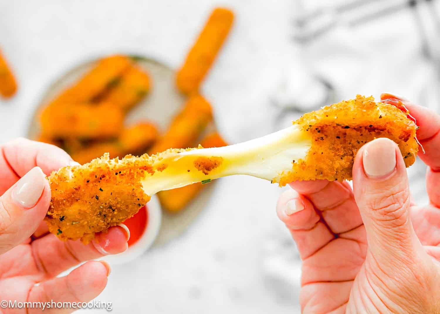 Close-up of hands pulling apart an Eggless Mozzarella Stick, with melted cheese stretching between the halves. A plate with more mozzarella sticks is blurred in the background.