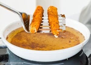 A slotted spatula lifts two Eggless Mozzarella Sticks out of a white frying pan filled with hot oil.