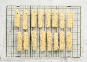 Fifteen Eggless Mozzarella Sticks arranged in rows on a cooling rack over a light-colored surface.