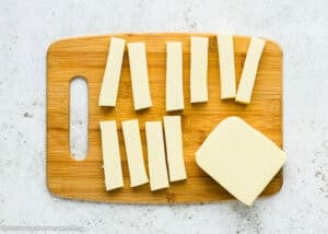 Eggless Mozzarella Sticks are served as rectangular slices of cheese arranged on a wooden cutting board, with a larger block of cheese beside them.