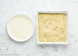 A round bowl of liquid batter sits next to a square dish filled with breadcrumbs, ready for making Eggless Mozzarella Sticks, on a white textured surface.