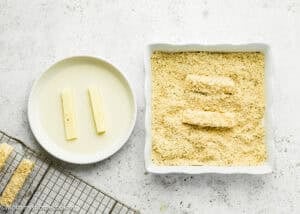Two Eggless Mozzarella Sticks are coated in a bowl, while three are covered in breadcrumbs in a square dish; a cooling rack with more sticks is nearby.
