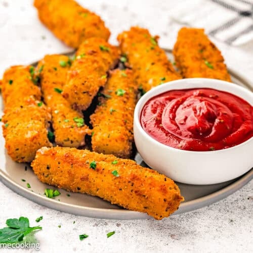 A plate of eggless mozzarella sticks garnished with herbs, served with a bowl of tomato dipping sauce on the side.