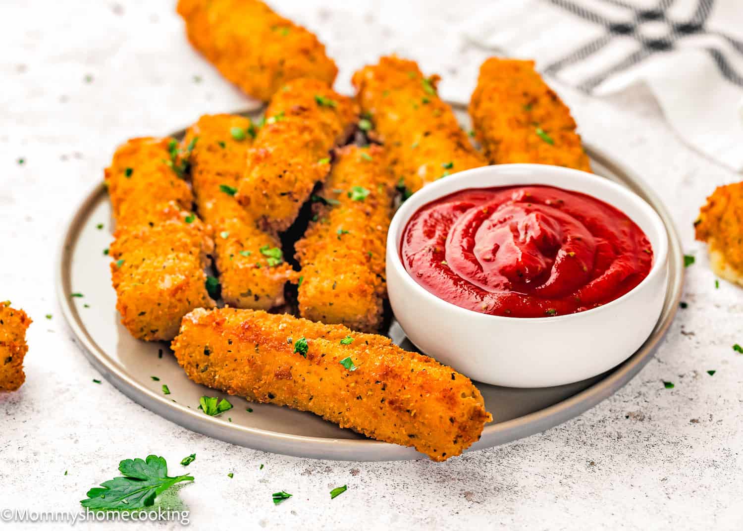 A plate of eggless mozzarella sticks garnished with herbs, served with a bowl of tomato dipping sauce on the side.