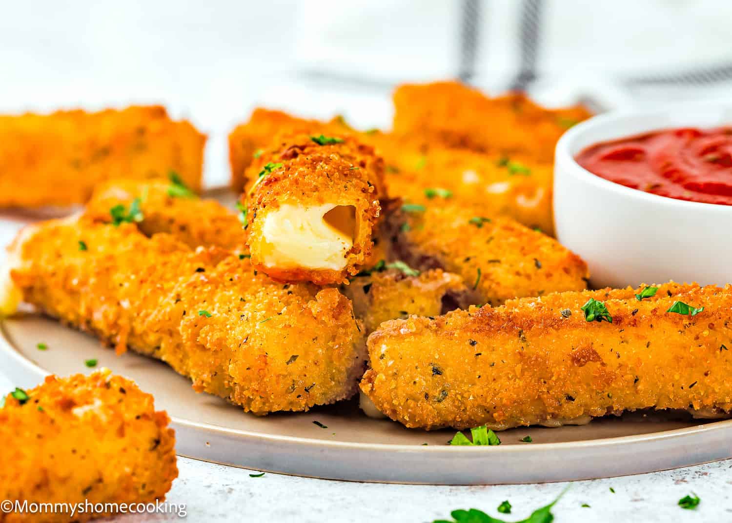 A plate of Eggless Mozzarella Sticks, with one broken open to reveal melted cheese inside, served alongside a bowl of marinara sauce.